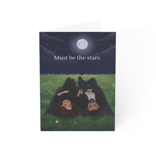 Greeting card with illustration of two people lying on grass under a moonlit sky, text 'Must be the stars'.